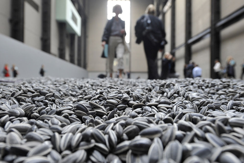 A photo from a low perspective showing sunflower seeds on the floor in the foreground, with out of focus people in the background.