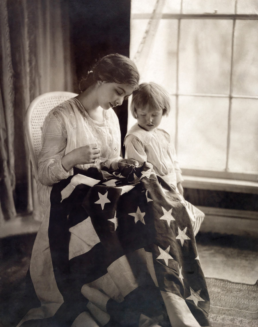 A child watches as a woman sews a star on a United States flag, 1917. Photograph by May Smith, National Geographic